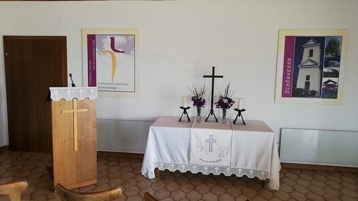 Ein bescheiden gestalteter Kapelleninnenraum mit Altar, Lesepult, Stühlen und religiösen Wandverzierungen.