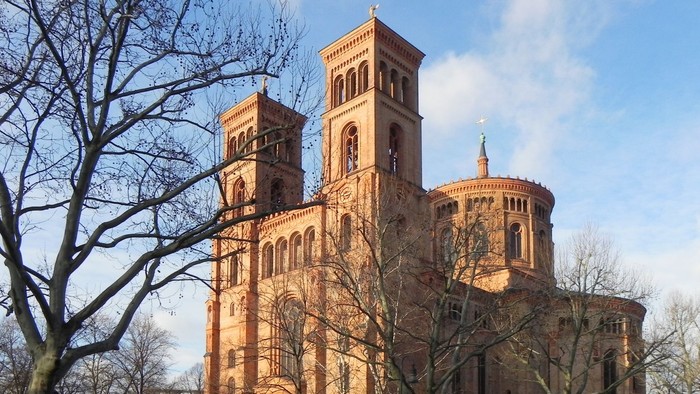 Historische Backsteinkirche mit Zwillingstürmen und rundem Chor, umgeben von kahlen Bäumen und parkenden Autos.