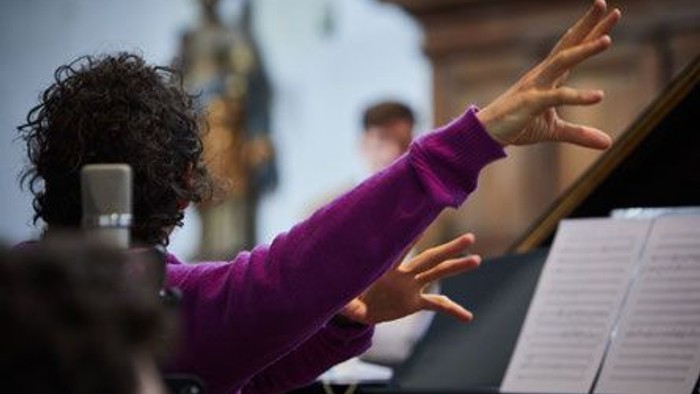Der Künstler Bartussek ist im fuchsia farbenen Pulli von hinten zu sehen, rechts ein Notenblatt und seine Arme sind zum Dirigieren erhoben