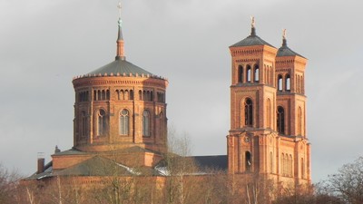 Zwei Türme einer Kirche mit spitzen Dächern und goldenen Akzenten, umgeben von Bäumen und einem bewölkten Himmel