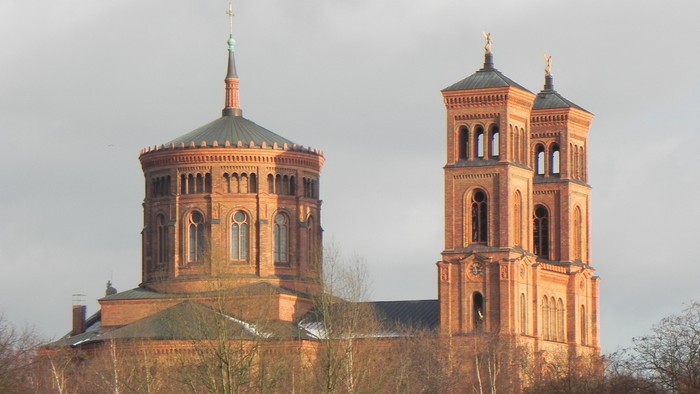 Zwei Türme einer Kirche mit spitzen Dächern und goldenen Akzenten, umgeben von Bäumen und einem bewölkten Himmel