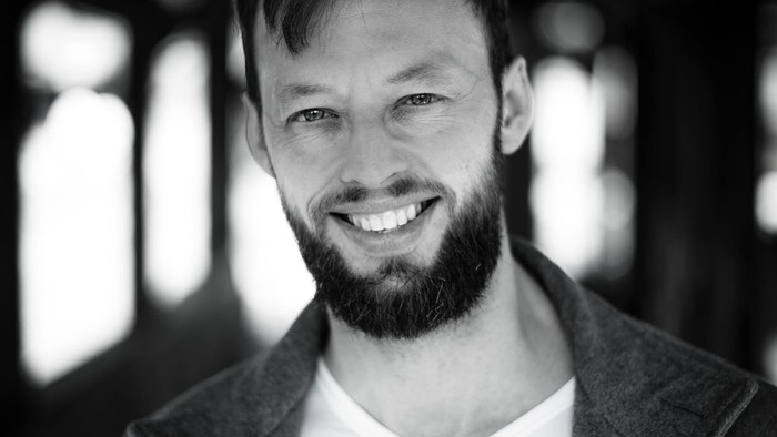 Mand med skæg, der smiler, iført en blazer og hvid skjorte i en svagt oplyst indendørs omgivelse.