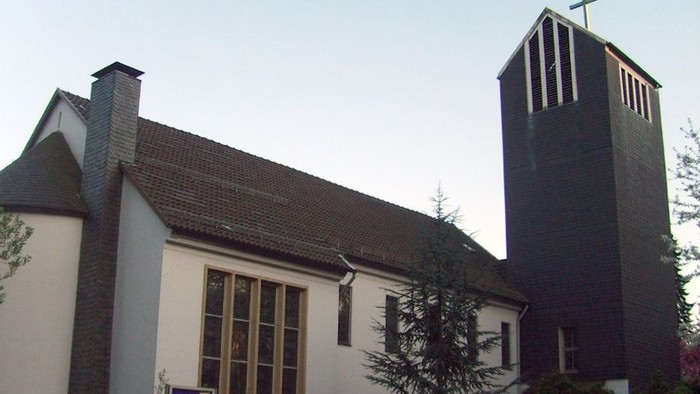 Moderne Kirche mit hohem Glockenturm und Kreuz, umgeben von Grünflächen unter klarem Himmel.
