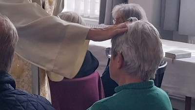 Ein Geistlicher segnet während einer religiösen Zeremonie den Kopf einer älteren Person, während andere in der Nähe sitzen.