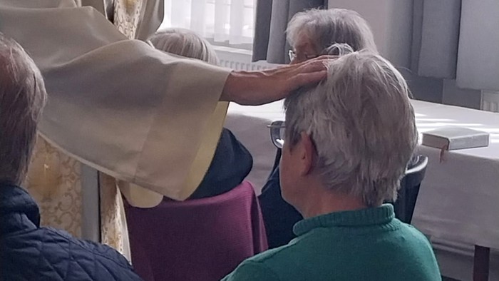 Ein Geistlicher segnet während einer religiösen Zeremonie den Kopf einer älteren Person, während andere in der Nähe sitzen.