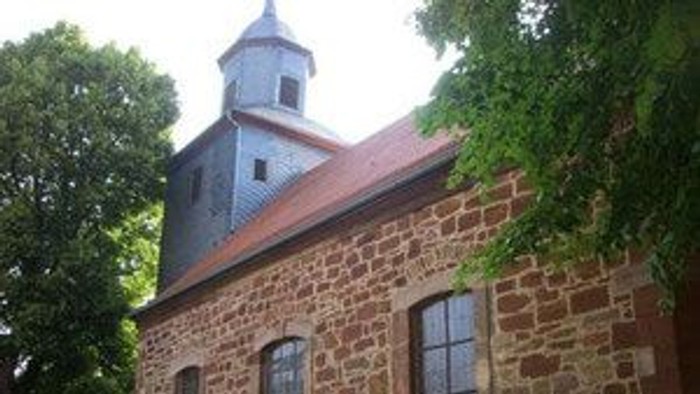 Historische Steinkirche mit Glockenturm und Bogenfenstern, umgeben von Grün.