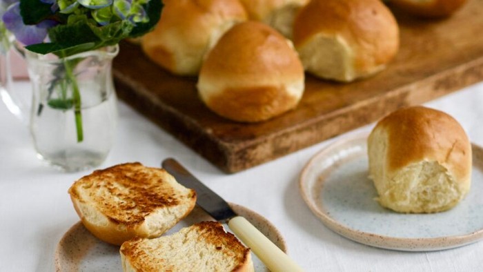 Friskristede boller med smør, anrettet på et træbræt og en tallerken ved siden af en lille vase med blomster.