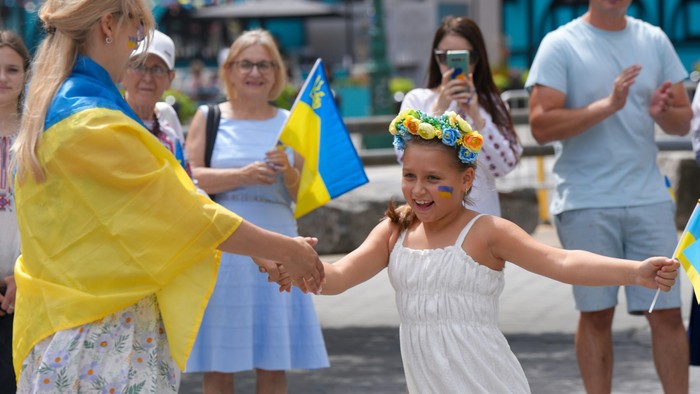 En ung pige danser med en voksen, mens de holder hinanden i hænderne, omgivet af mennesker, der vinker med blå og gule flag udendørs.