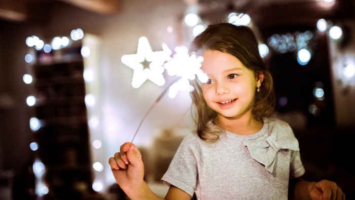 Et glad barn holder en glødende stjerneformet sparkler og smiler strålende indendørs med festlige lys i baggrunden.