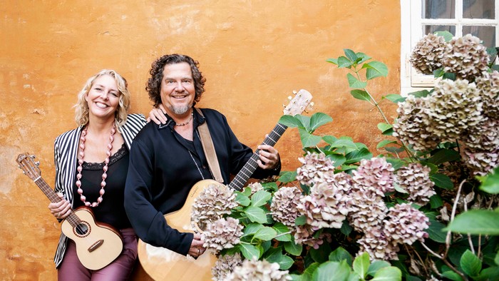 To musikere med guitarer står sammen ved siden af en farverig, tekstureret mur og blomstrende planter.