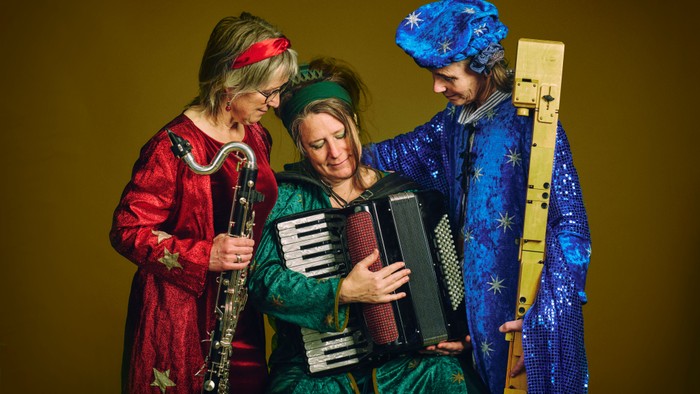 Tre musikere i farverige kostumer spiller på instrumenter på scenen under en festlig optræden.