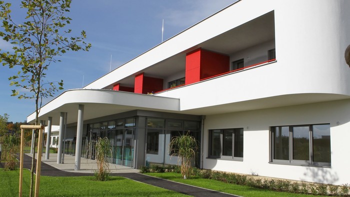 Modernes Gebäude mit roten Akzenten und großem Fensterbereich, umgeben von Grünfläche und Bäumen