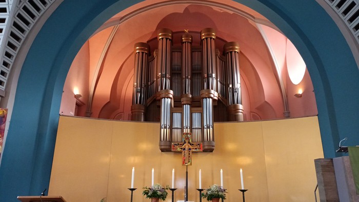 Elegante Kircheninnenausstattung mit einer großen Orgel über einem schlichten Altar mit Kerzen und Topfpflanzen.