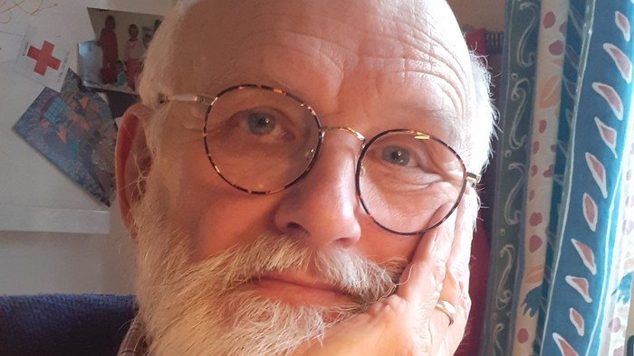 En mand med briller og skæg hviler hagen i hånden i en hyggelig indendørs indstilling.