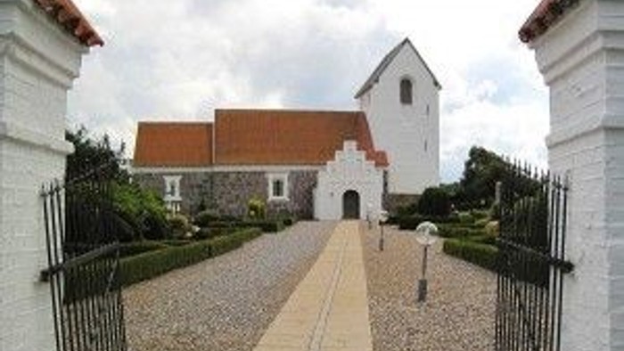 En smuk og lille kirke med røde tag og hvidt tårn, omgivet af en mur og en sti.