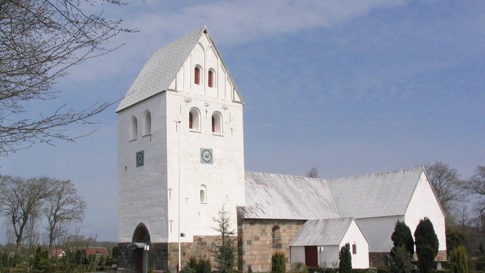 En hvid kirke med tårn og klokke, omgivet af træer og gravhøje
