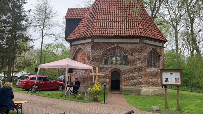 Ein kleines Backsteingebäude mit rotem Dach und einem Zelt davor, umgeben von Bäumen und Menschen.
