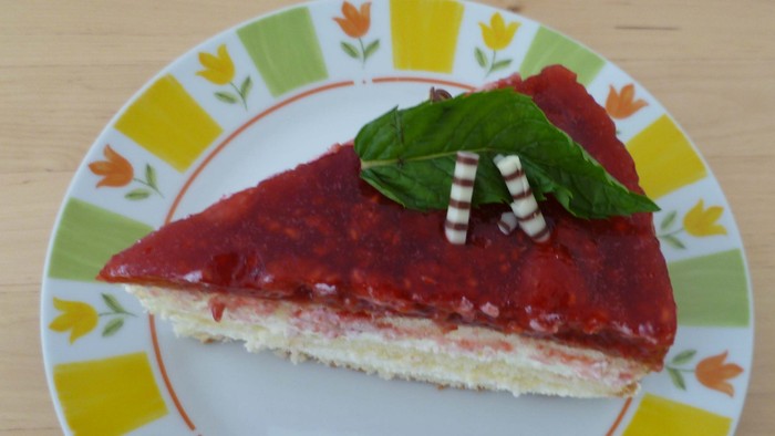 Ein Stück cremiger Käsekuchen mit Erdbeerglasur, Minzblatt und Crackern auf einem dekorativen Teller.