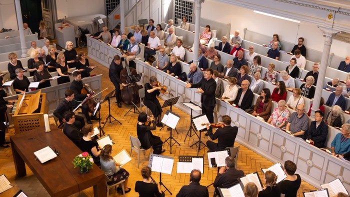 Ein Orchester probt in einem großen Saal vor einem Publikum.