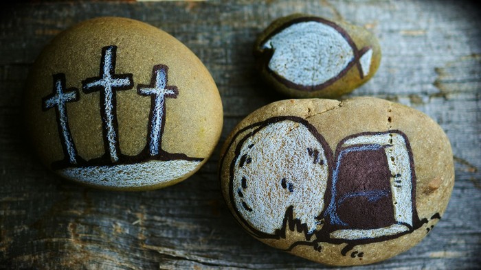 Verzierte glatte Steine, die Kreuze, Fische und ein leeres Grab auf einer Holzoberfläche symbolisieren.