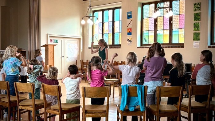 Die Kinder versammeln sich aufmerksam in einem klassenzimmerähnlichen Raum und hören einem Lehrer an einem hölzernen Pult zu.