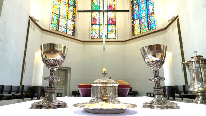 Elegante Kirchenaltar-Anordnung mit verzierten Kelchen, einer Kerze und farbenfrohen Buntglasfenstern im Hintergrund.