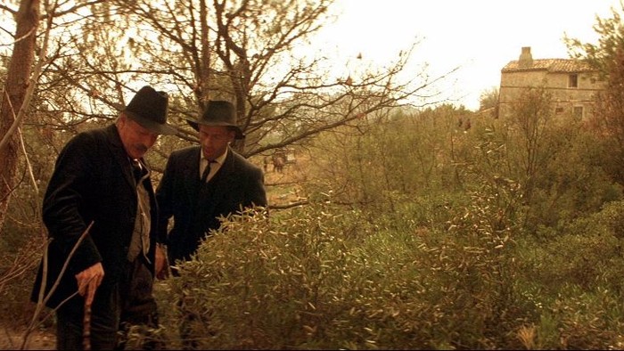 To mænd i vintage-tøj går gennem et tæt, overgroet landskab nær en fjern bygning.