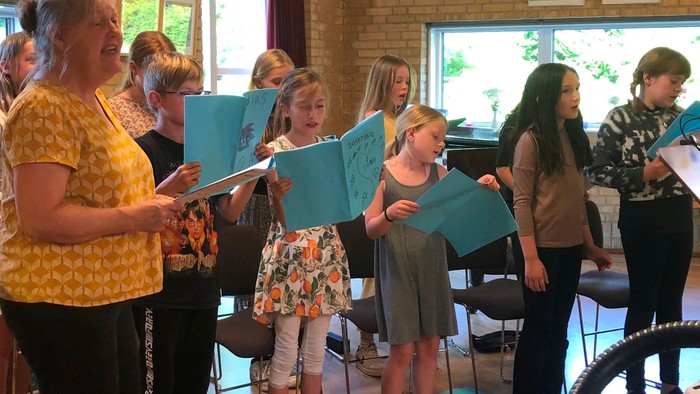 En gruppe af børn og voksne står i en sangkveld, hvor de holder blå sanghefter og synger.