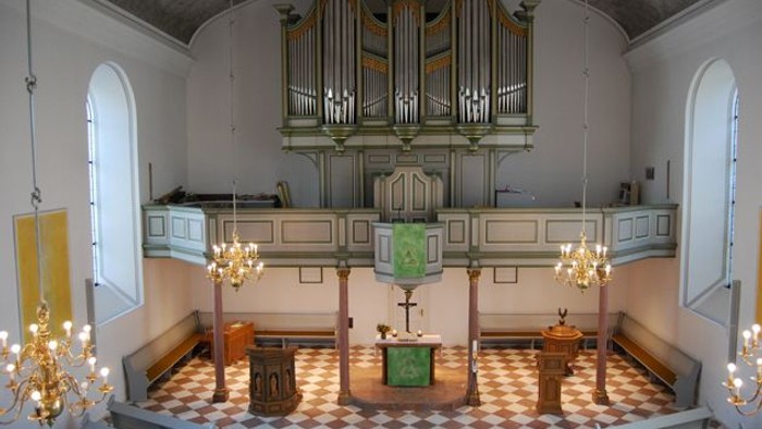 Elegante Kircheninnenausstattung mit einer großen Orgel, einem Altar und dekorativen Kronleuchtern auf einem karierten Boden.
