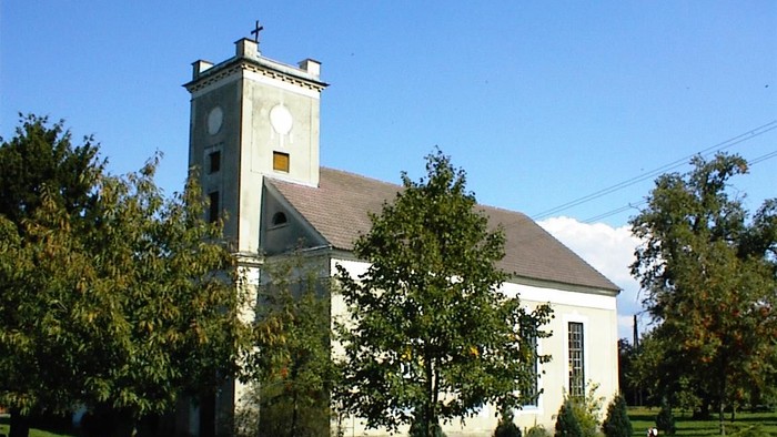 Weiße Kirche mit hohem Turm und Uhr, umgeben von Bäumen und Gras.