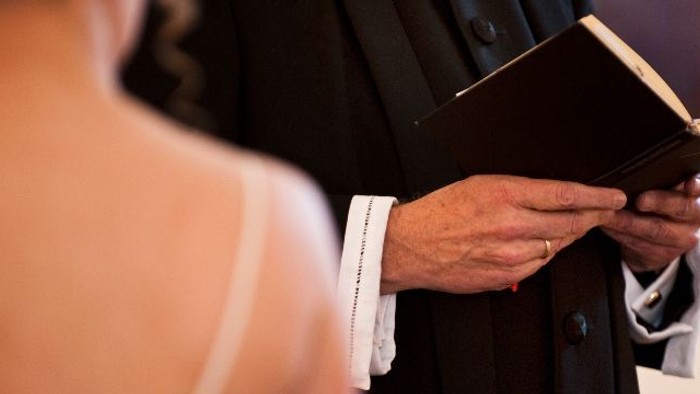 En person i formelt tøj læser op fra en åben bog under en ceremoni, med en anden person i en hvid kjole ved siden af.