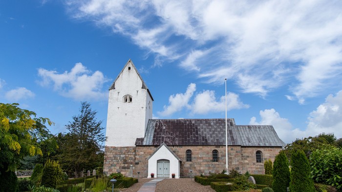 En smuk kirke med tårn og flagstang foran, omgivet af grønne planter og træer under et blødt blå himmel.