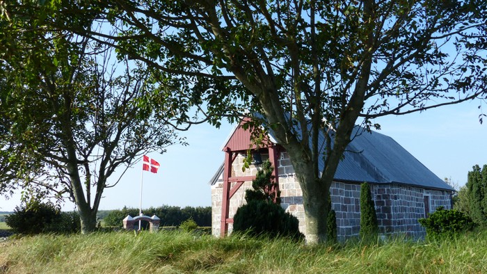 En lille rød kapel med blå tag og en dansk flag ved en græslig plads med træer og sten.