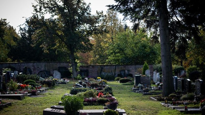 Friedhof mit Gräbern, Blumen und Bänken, umgeben von Bäumen.