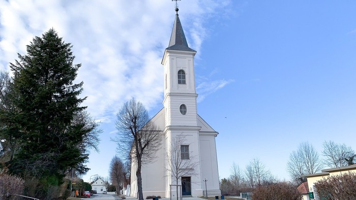 Weiße Kirche mit hohem Turm und Uhr, umgeben von Bäumen und Himmel.