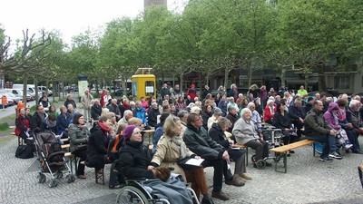 Große Menschenmenge sitzt im Freien, viele im Rollstuhl, vor einem gelben Bus