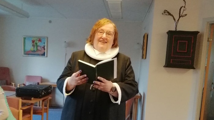 En kvinde i mørkt tøj og hvid krage står og læser fra en bog i et rum med en anden kvinde i baggrunden.
