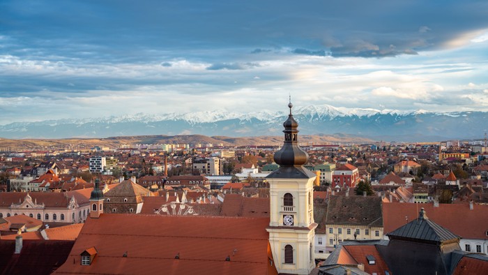 Panoramablick auf eine europäische Stadt mit rotgedeckten Gebäuden und schneebedeckten Bergen im Hintergrund.