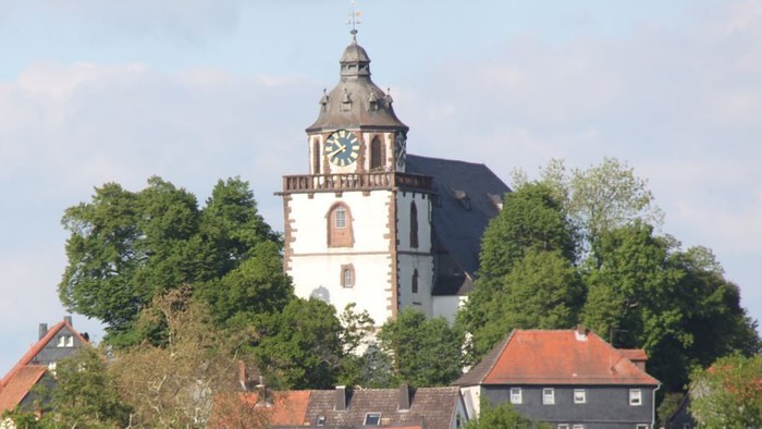 Historischer Kirchturm mit Uhr thront markant über einem grünen, üppigen Hügeldorf.