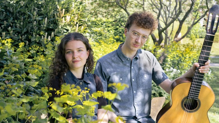 To personer sidder udendørs omgivet af frodig grønne omgivelser, hvor den ene holder en lys klassisk guitar.