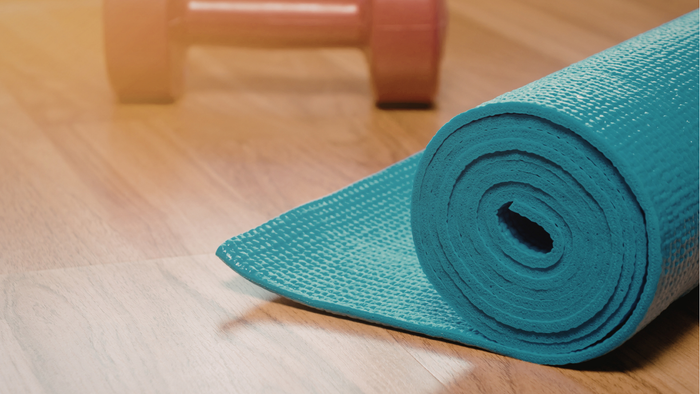 Eine gerollte türkisfarbene Yogamatte liegt neben roten Kurzhanteln auf einem Holzboden.