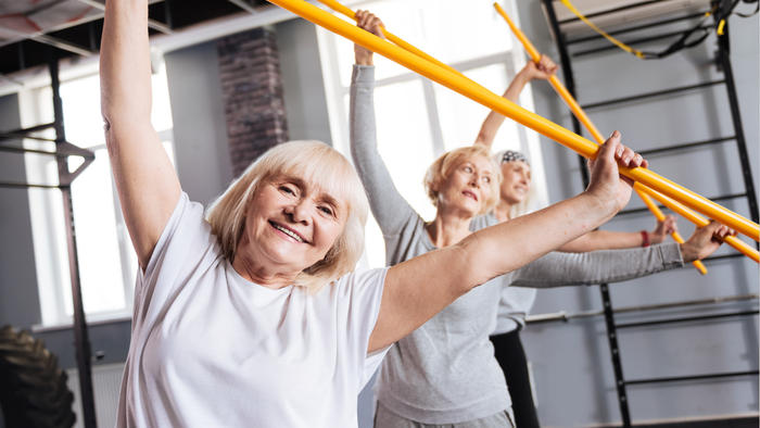 Drei ältere Frauen trainieren fröhlich mit gelben Widerstandsbändern in einem Fitnessstudio.