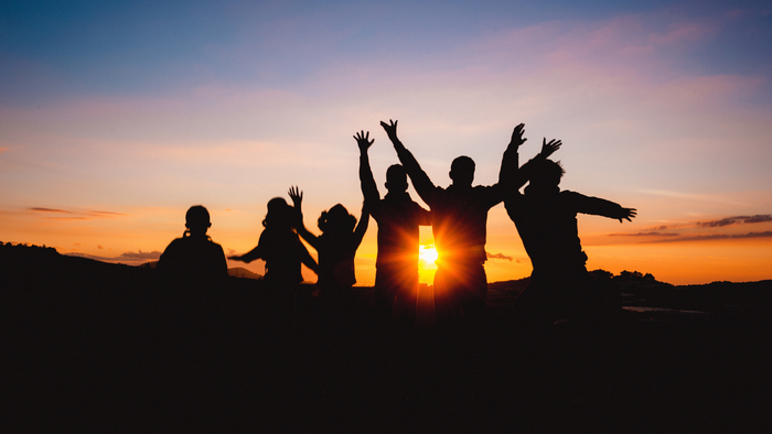 Gruppe feiert fröhlich bei Sonnenuntergang mit erhobenen Armen vor einem leuchtenden Himmel.