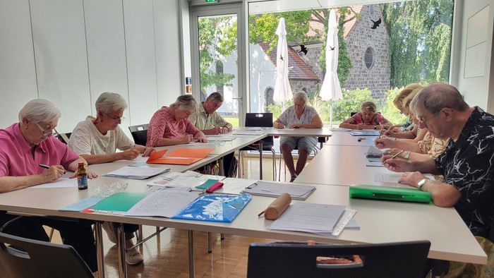 Seniorengruppe sitzt an Tischen und arbeitet in einem hellen Raum mit großen Fenstern.