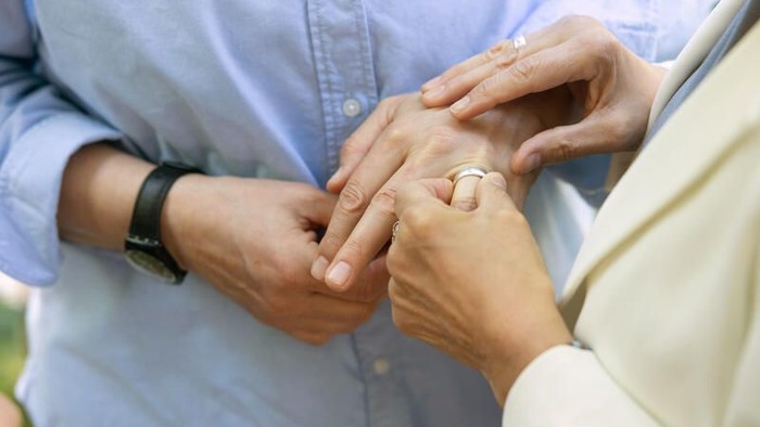 Zwei Menschen, die sich an den Händen halten, einer trägt einen Ring, und teilen einen zärtlichen Moment im Freien.