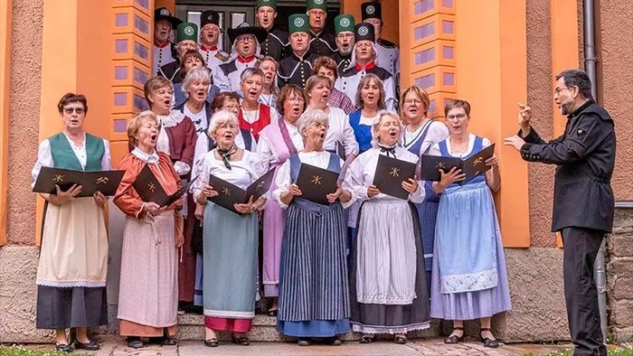 Ein traditioneller Chor in Tracht tritt vor einem Gebäude auf, während ein Dirigent sie leitet.