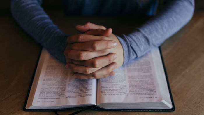 Eine Person mit gefalteten Händen im Gebet über einem offenen Buch, wahrscheinlich einer Bibel, auf einer hölzernen Oberfläche.