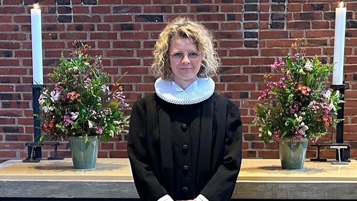 Kvinde i sort kapok med hvid krage står foran alter med blomsterarrangementer og lysestake.