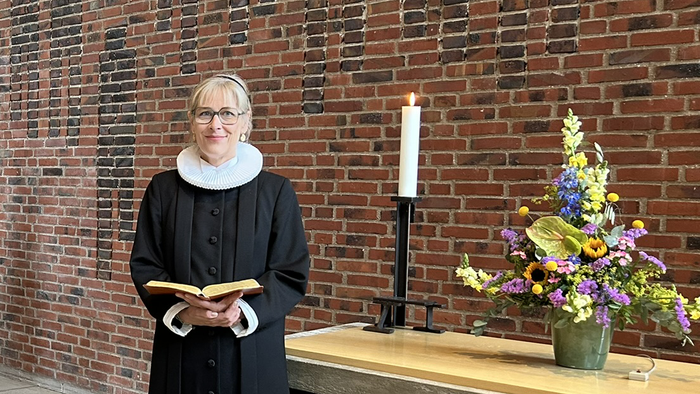 En kvinde i svart kapittel holder en bog og står foran en mur af røde mursten.