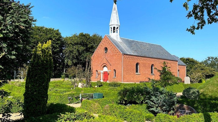 En rød murstenskirke med spir med omgivelser af grønne planter og træer